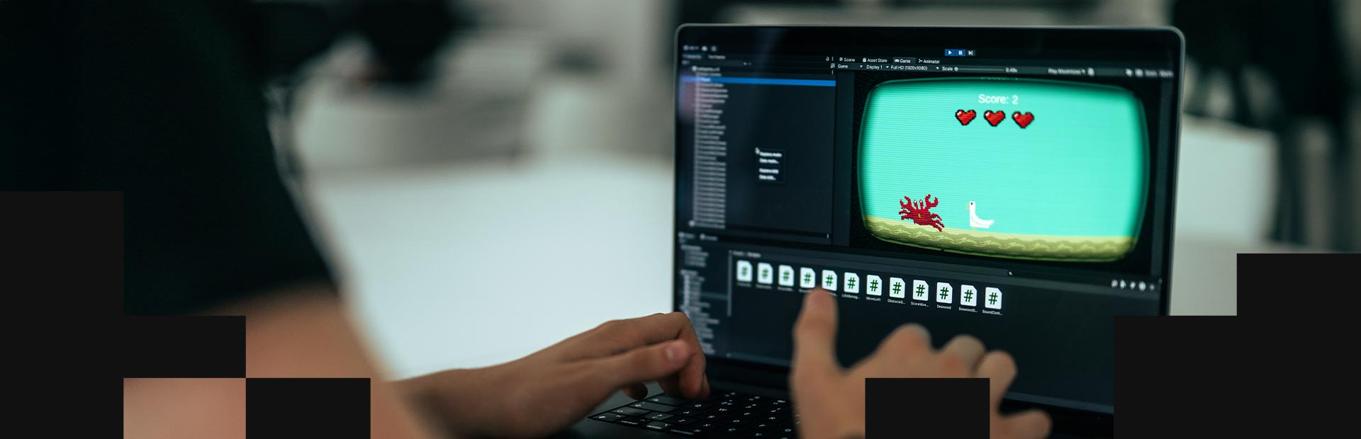 En person arbetar vid en bärbar dator med ett spelutvecklingsprogram öppet. På skärmen syns ett retroinspirerat plattformsspel med en krabba, en fågel och tre hjärtan som indikerar liv.