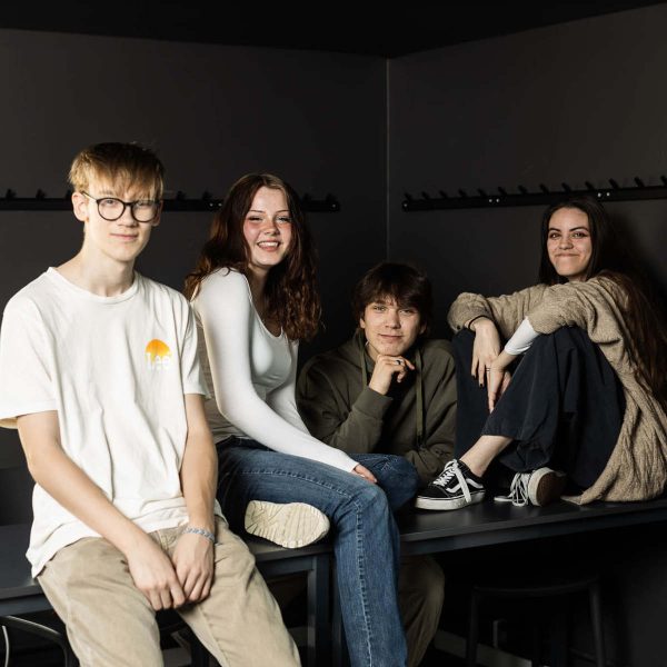Fyra elever hänger tillsammans i ett svart klassrum och tittar in i kameran.