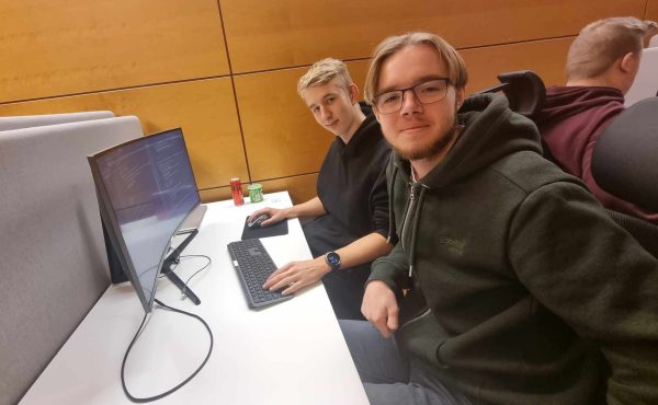 Två elever sitter vid en skärm med kod och tittar in i kameran.